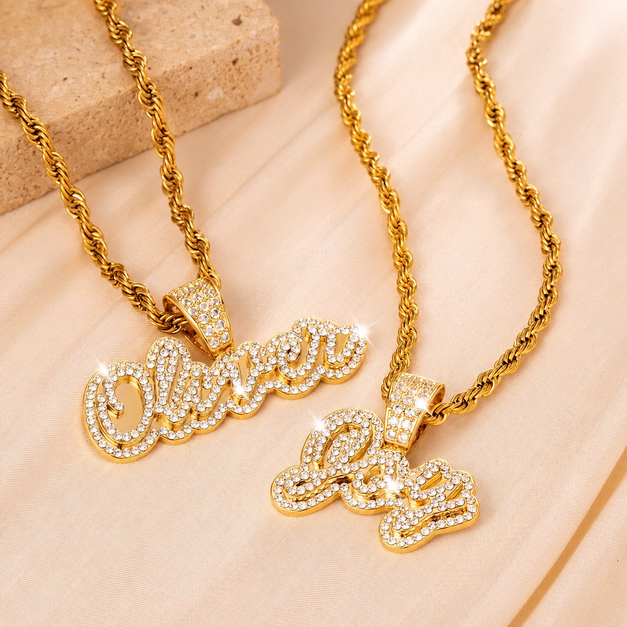 Two gold Crystal Cursive Name Necklaces with sparkling custom name pendants, crystal-studded bails, and rope chains displayed on cream satin